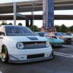 Une Honda e blanche lourdement modifiée (posée au sol, grosses jantes) au premier plan d'un rassemblement automobile à Daikoku Parking Area dans Forza Horizon 6. En arrière-plan, une Mazda MX-5 Miata modifiée avec un moteur exposé et d'autres voitures JDM sous des viaducs d'autoroute.