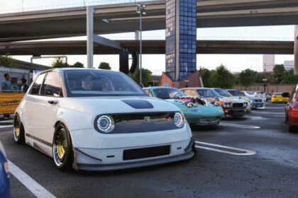 Une Honda e blanche lourdement modifiée (posée au sol, grosses jantes) au premier plan d'un rassemblement automobile à Daikoku Parking Area dans Forza Horizon 6. En arrière-plan, une Mazda MX-5 Miata modifiée avec un moteur exposé et d'autres voitures JDM sous des viaducs d'autoroute.