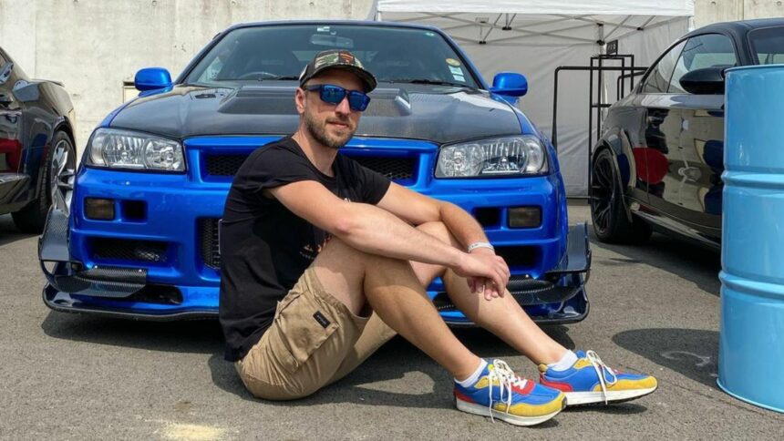 Victor Diakov assis devant une Nissan Skyline R34 GT-R bleue en train de recalculer ses mauvais choix de vie de 2013.