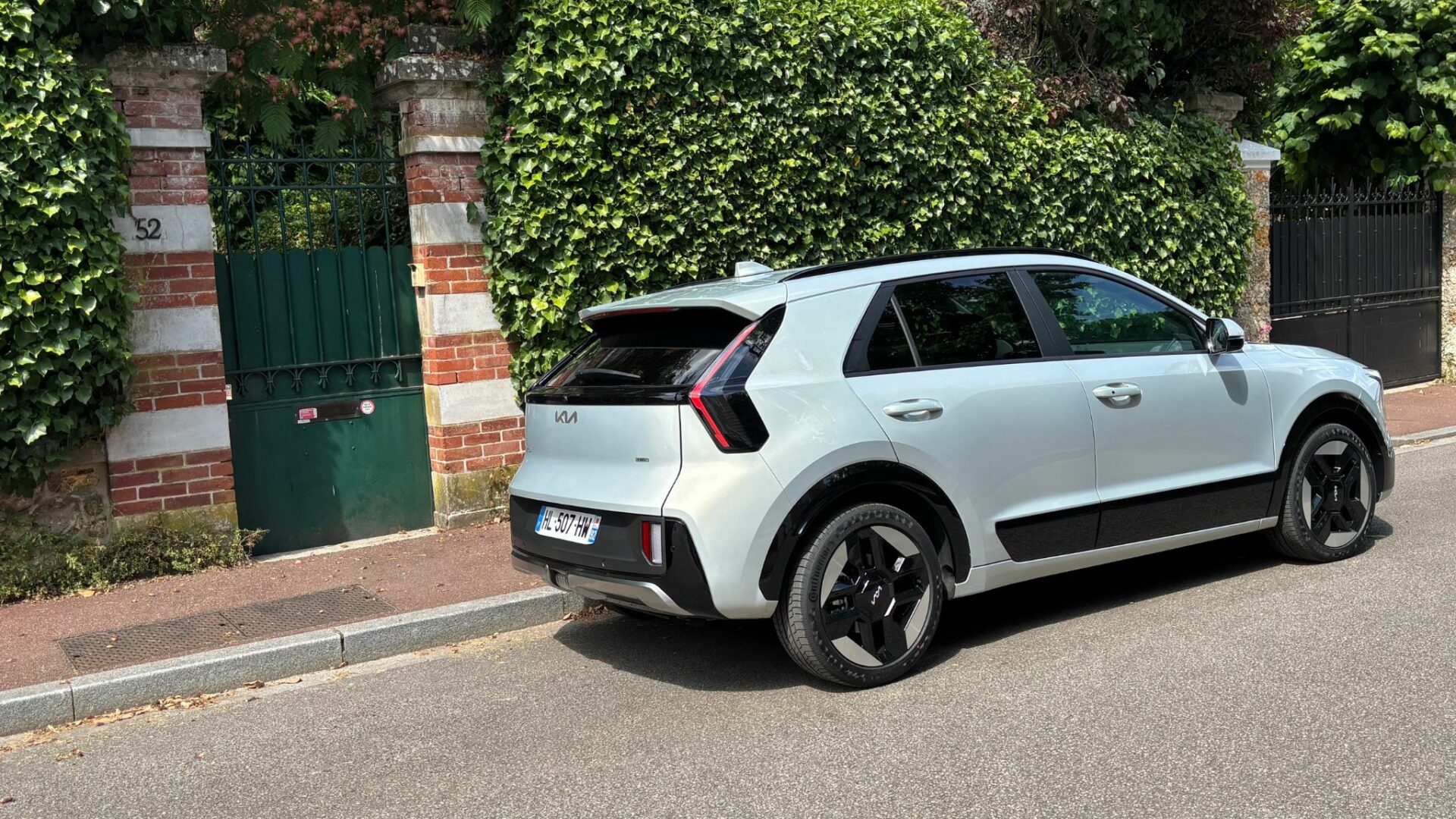 Vue de trois-quarts arrière du Kia Niro Hybride 2026 Gris Ivoire garé le long d'un trottoir devant un portail vert