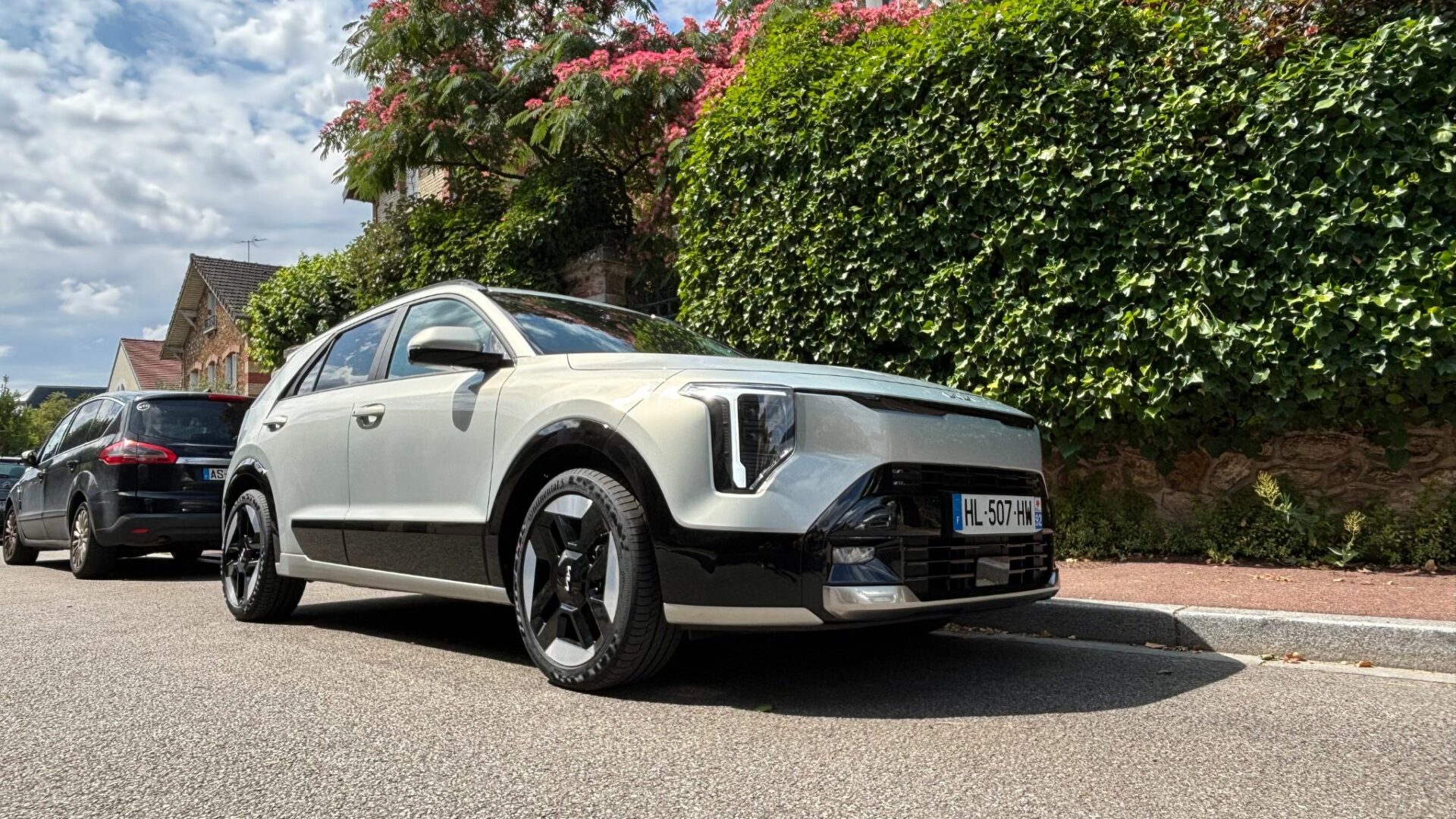 Vue en contre-plongée du Kia Niro Hybride 2026 Gris Ivoire stationné devant un mur végétal