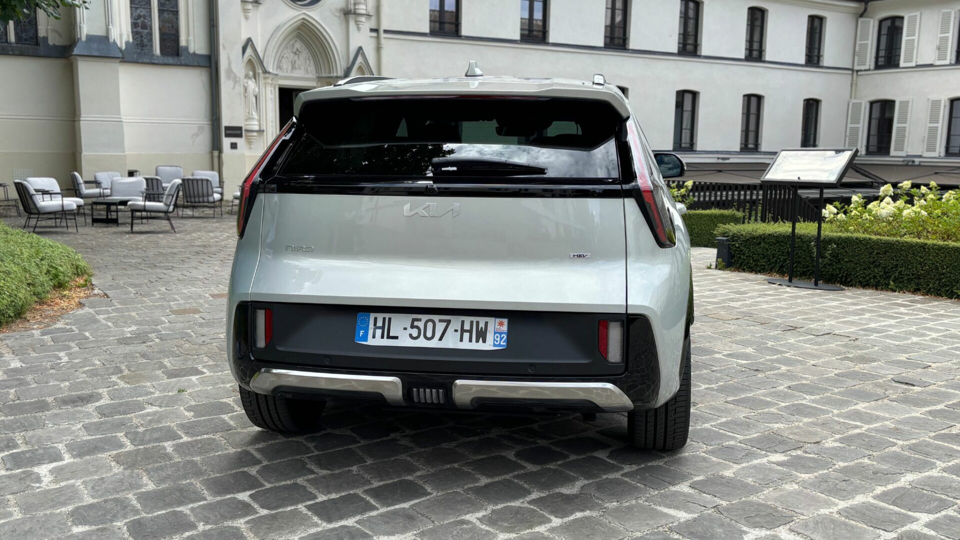 Vue arrière centrée du Kia Niro Hybride 2026 en teinte Gris Ivoire stationné sur un sol en pavés devant un bâtiment historique