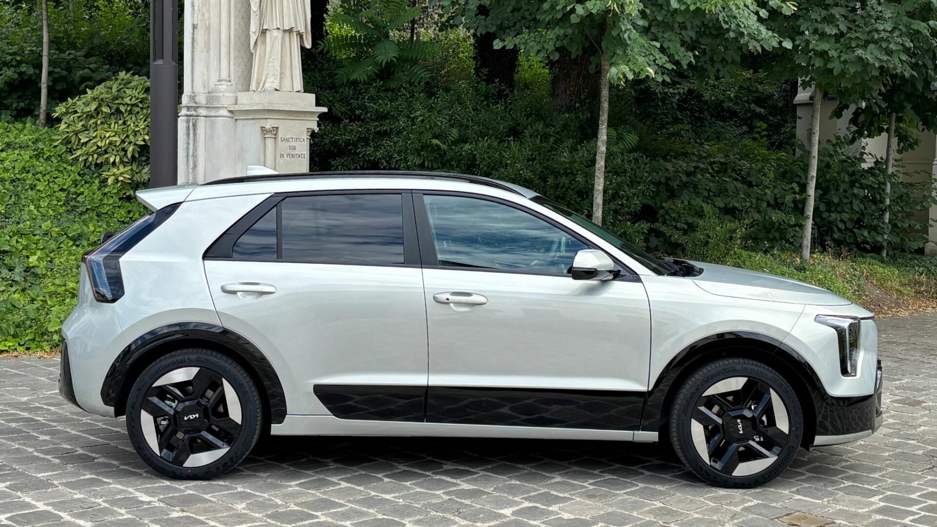 Vue de profil du Kia Niro Hybride 2026 en teinte Gris Ivoire stationné sur un sol pavé devant une statue et un cadre verdoyant