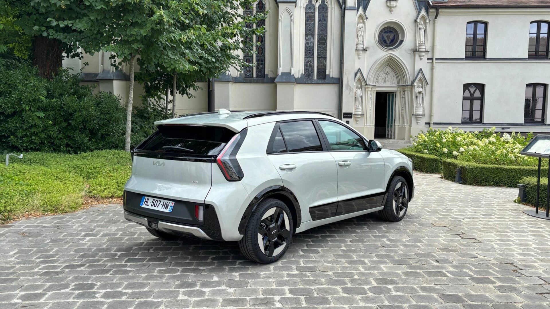 Kia Niro Hybride 2026 couleur Gris Ivoire stationné de trois-quarts arrière sur une cour pavée devant un bâtiment de style néogothique