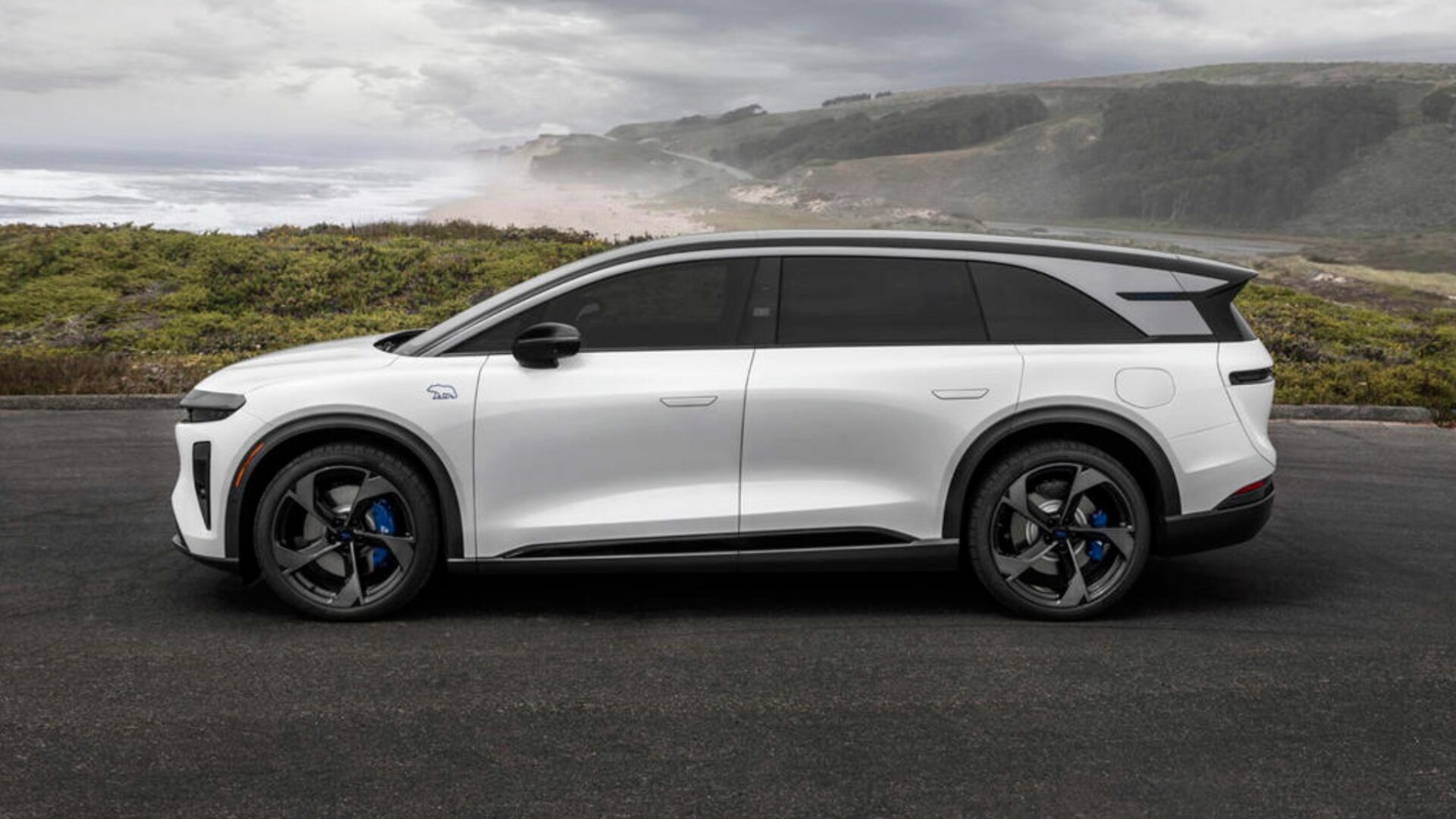 Vue de profil complète du SUV électrique blanc Lucid Gravity GT-S stationné au bord de l'océan avec l'ours bleu Sapphire sur l'aile avant