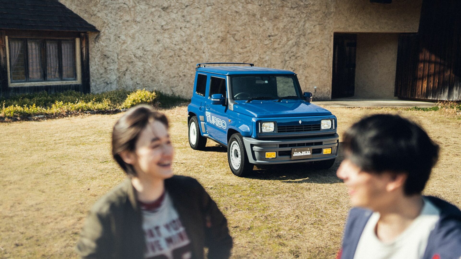 Suzuki Jimny R5 bleu de DAMD avec sticker Non Turbo sur le flanc, garé sur l'herbe derrière deux personnes floues
