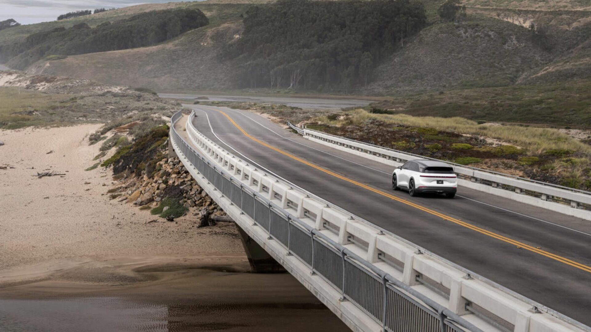 Vue de trois-quarts arrière du SUV électrique Lucid Gravity GT-S blanc roulant sur un pont au bord de la côte pacifique