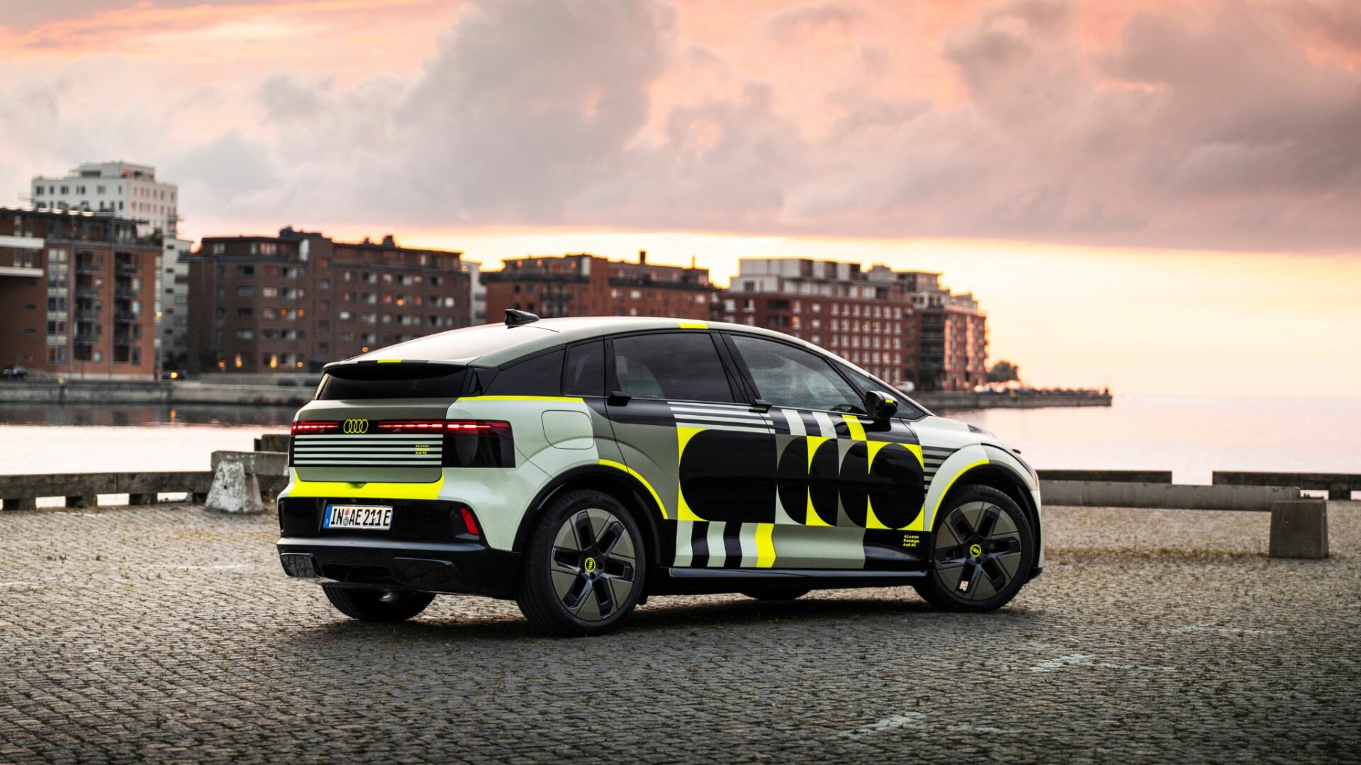 Audi A2 e-tron prototype camouflé stationné de trois-quarts arrière sur un quai pavé en bord de mer au coucher du soleil