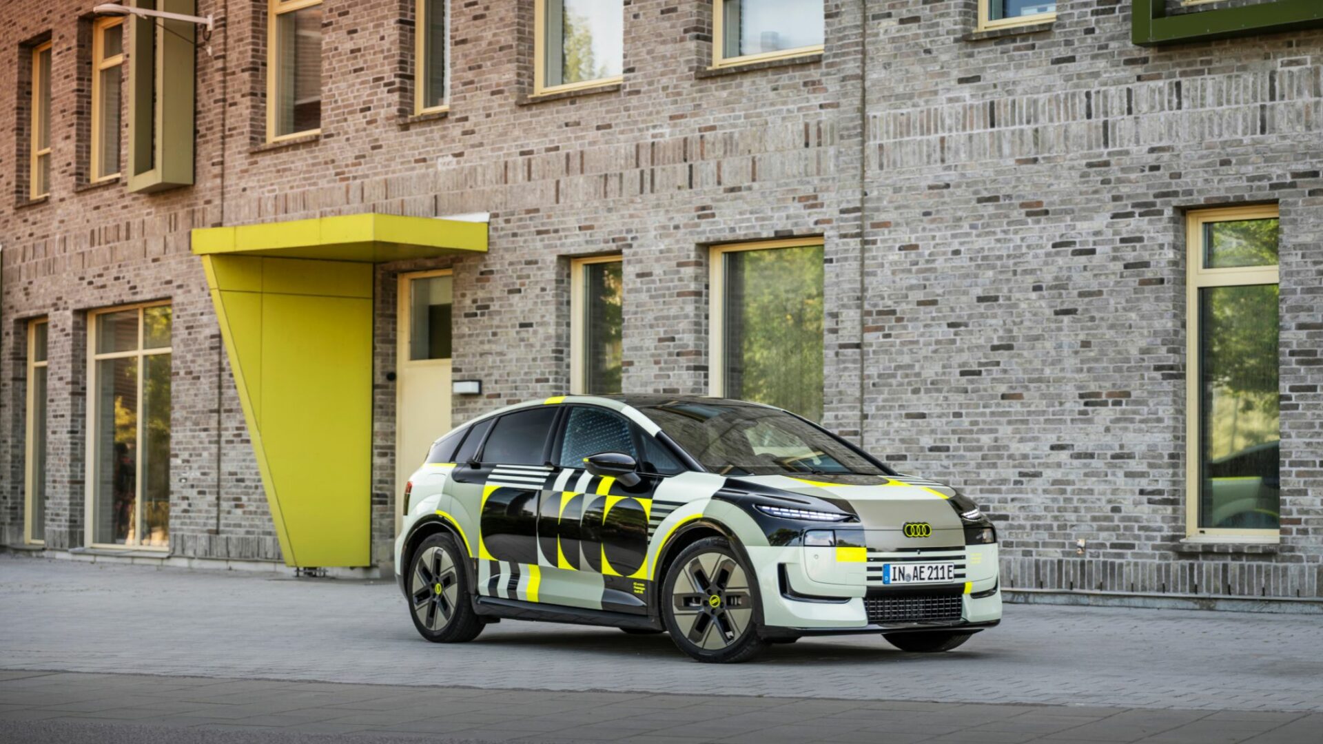 Vue d'ensemble de trois-quarts face du prototype Audi A2 e-tron camouflé stationné devant un immeuble moderne en briques