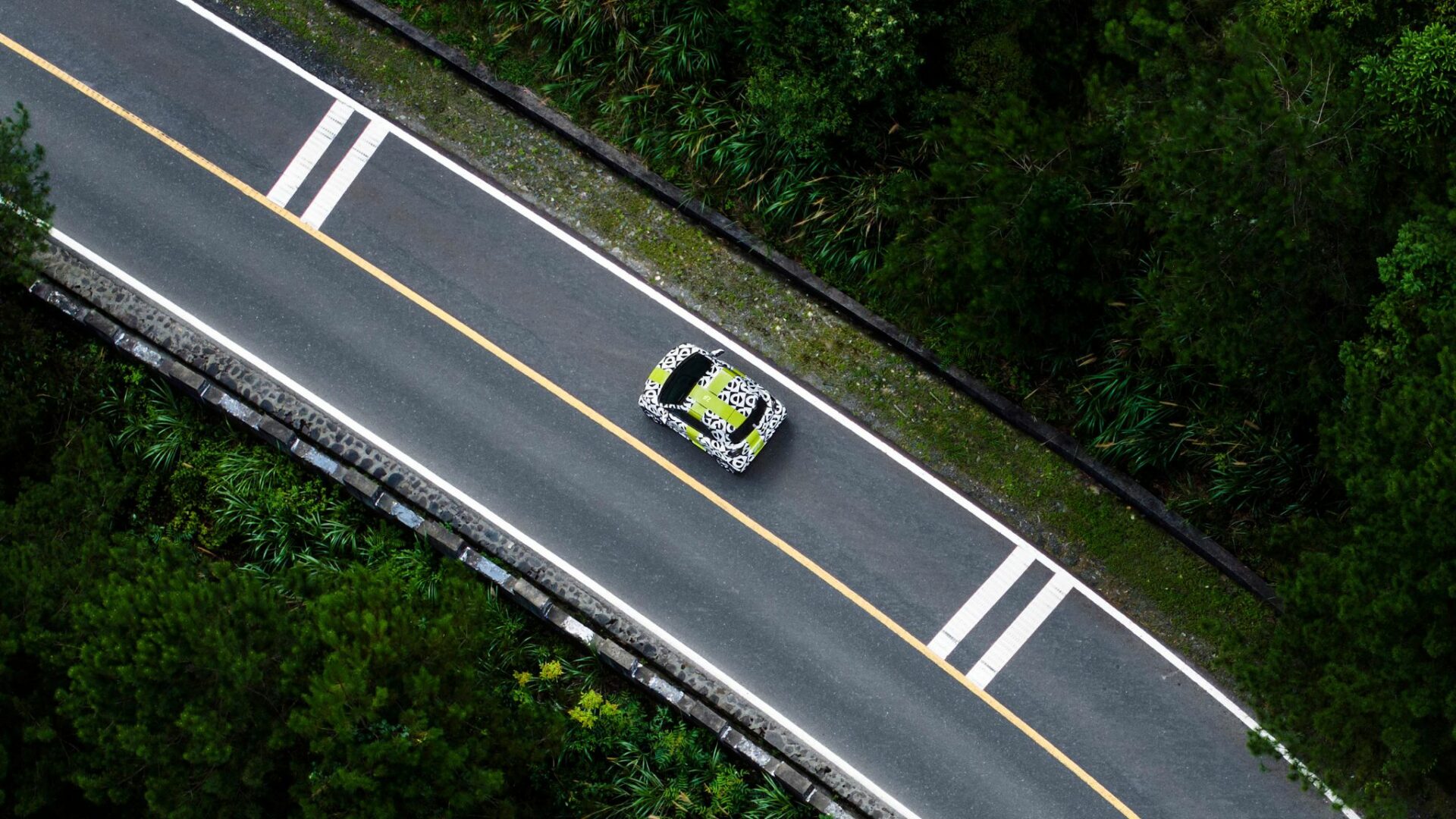 Vue aérienne plongeante en drone d'un prototype camouflé de Smart #2 électrique roulant sur une route à travers la forêt