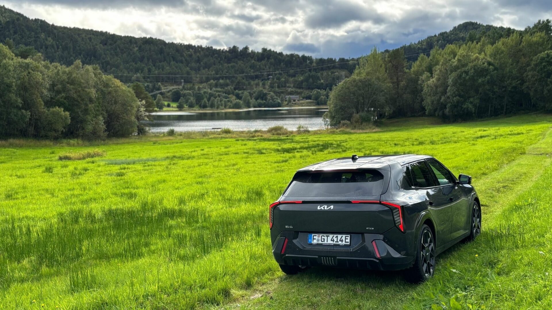 Vue de trois quarts arrière de la nouvelle Kia EV4 GT grise stationnée dans un grand champ verdoyant face à un lac et des collines boisées en Norvège