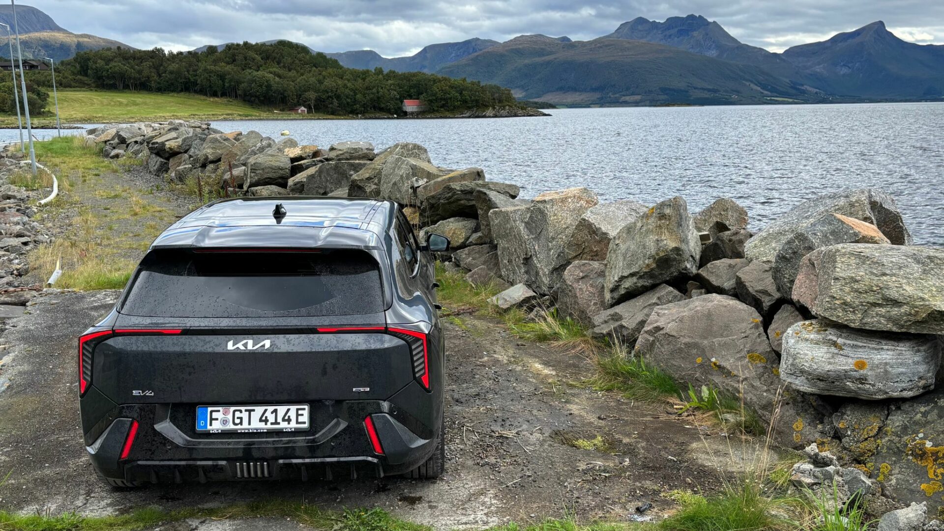 Vue arrière en plongée de la nouvelle Kia EV4 GT grise stationnée sur un chemin de pierres longeant l'eau face aux montagnes en Norvège.