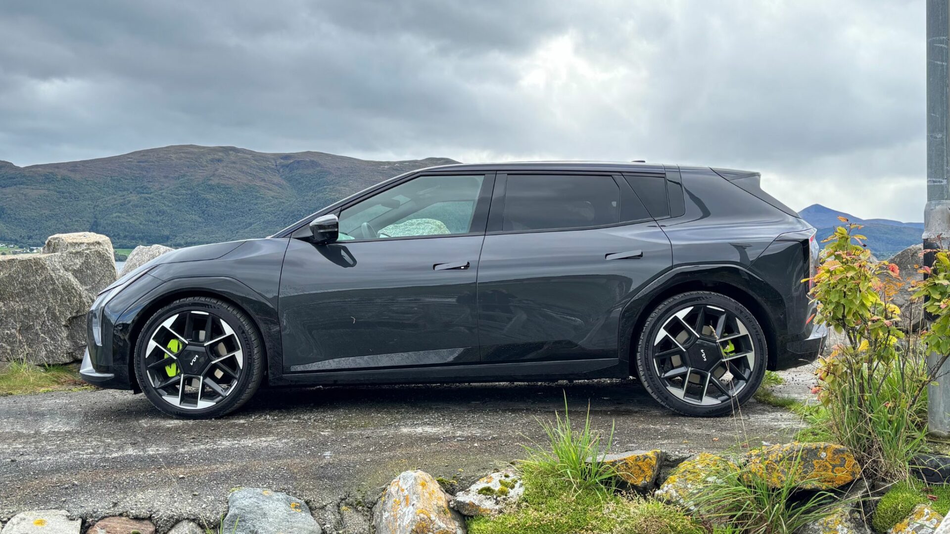 Vue de profil latéral de la nouvelle Kia EV4 GT grise stationnée sur un muret côtier face aux montagnes et aux nuages en Norvège.