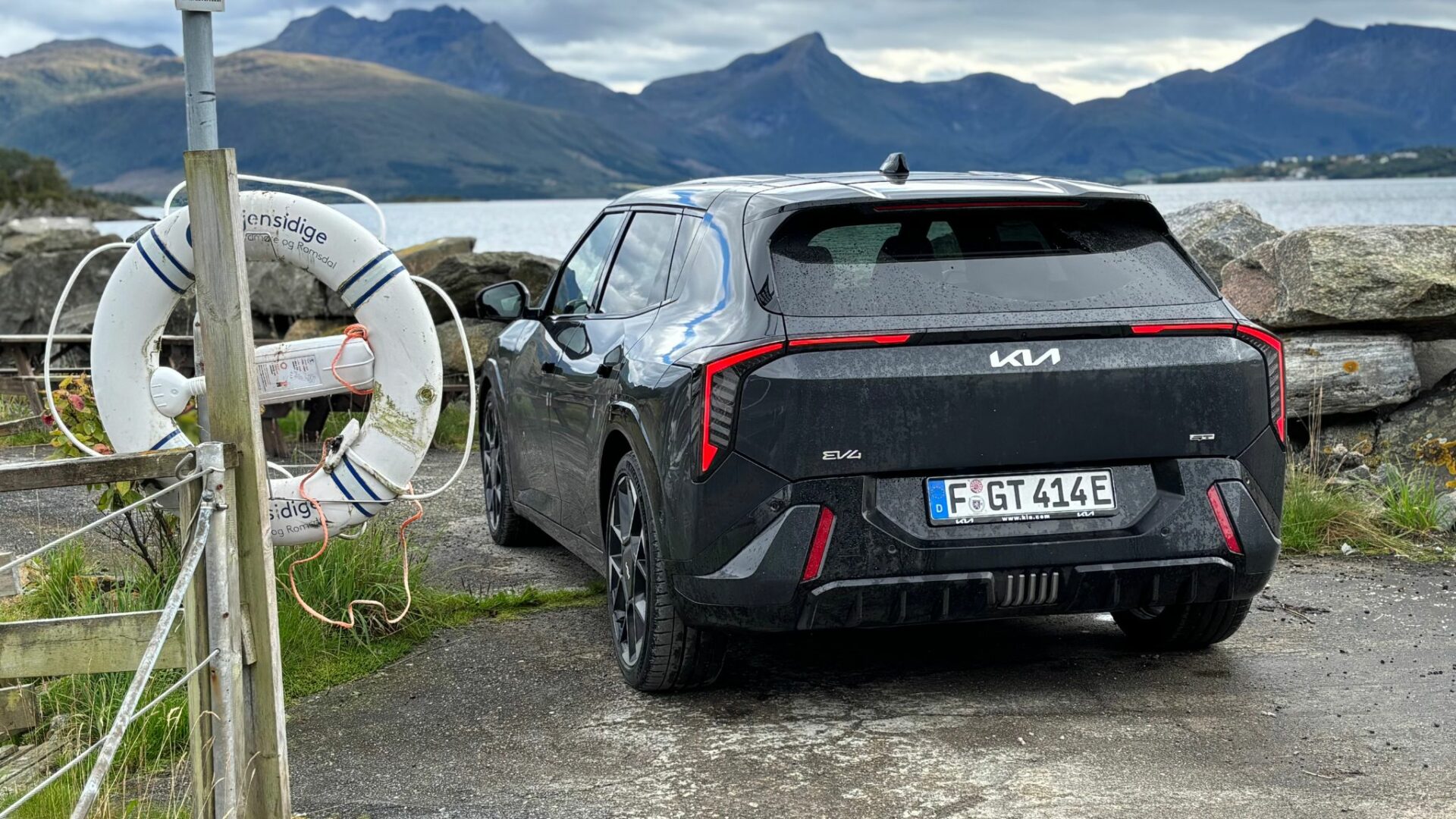 Vue de trois quarts arrière de la nouvelle Kia EV4 GT grise stationnée sur un quai près d'une bouée de sauvetage et d'un fjord entouré de montagnes en Norvège.