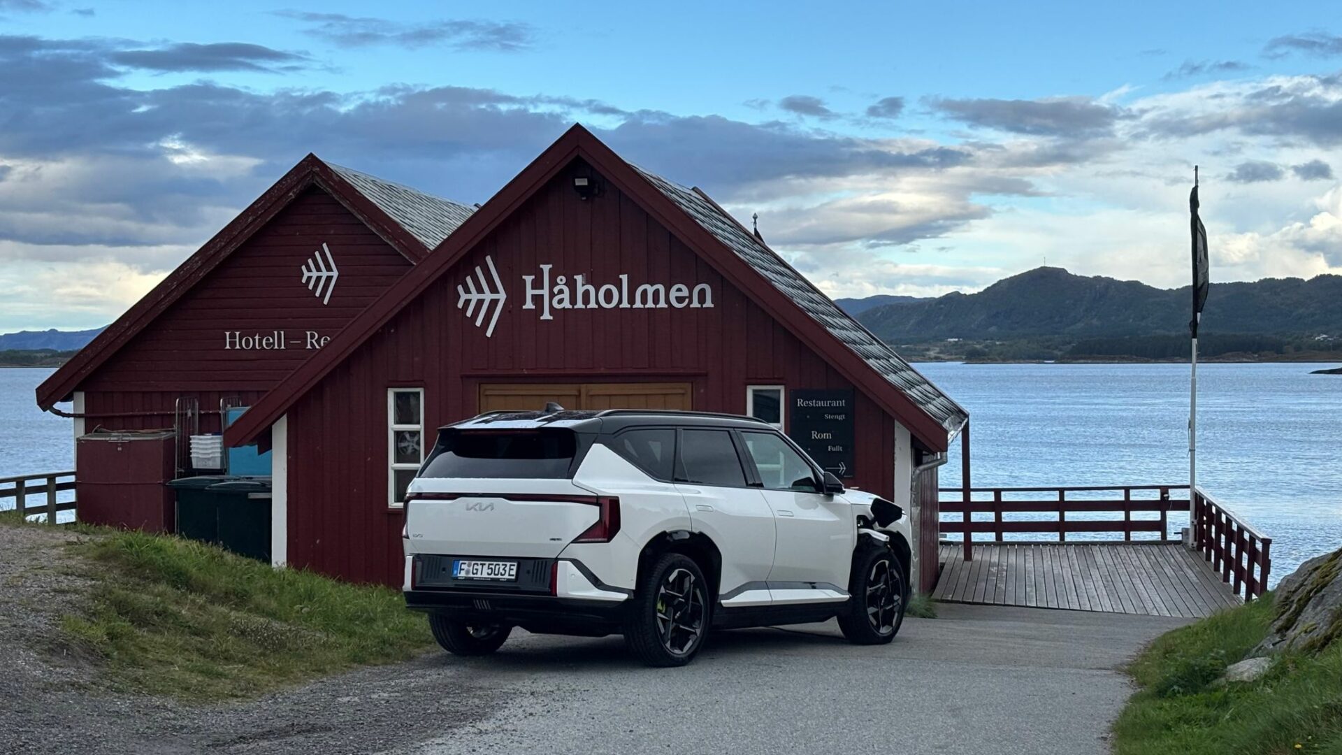 Vue de trois quarts arrière de la nouvelle Kia EV5 GT blanche stationnée au bord de l'eau devant un bâtiment en bois traditionnel, mettant en valeur son design de C-SUV sportif de 306 ch.