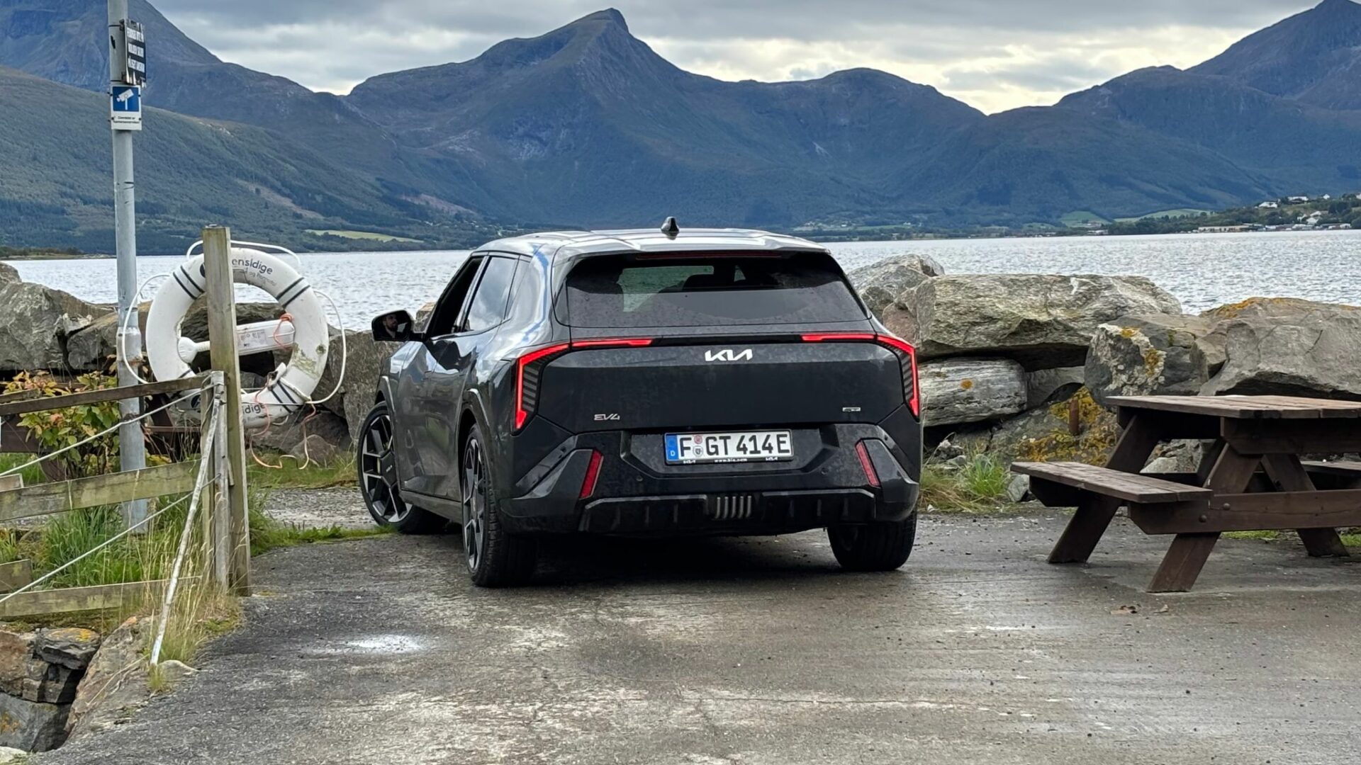 Vue de trois quarts arrière de la nouvelle Kia EV4 GT grise stationnée sur une aire de repos côtière avec une table en bois, une bouée de sauvetage et des montagnes en Norvège.