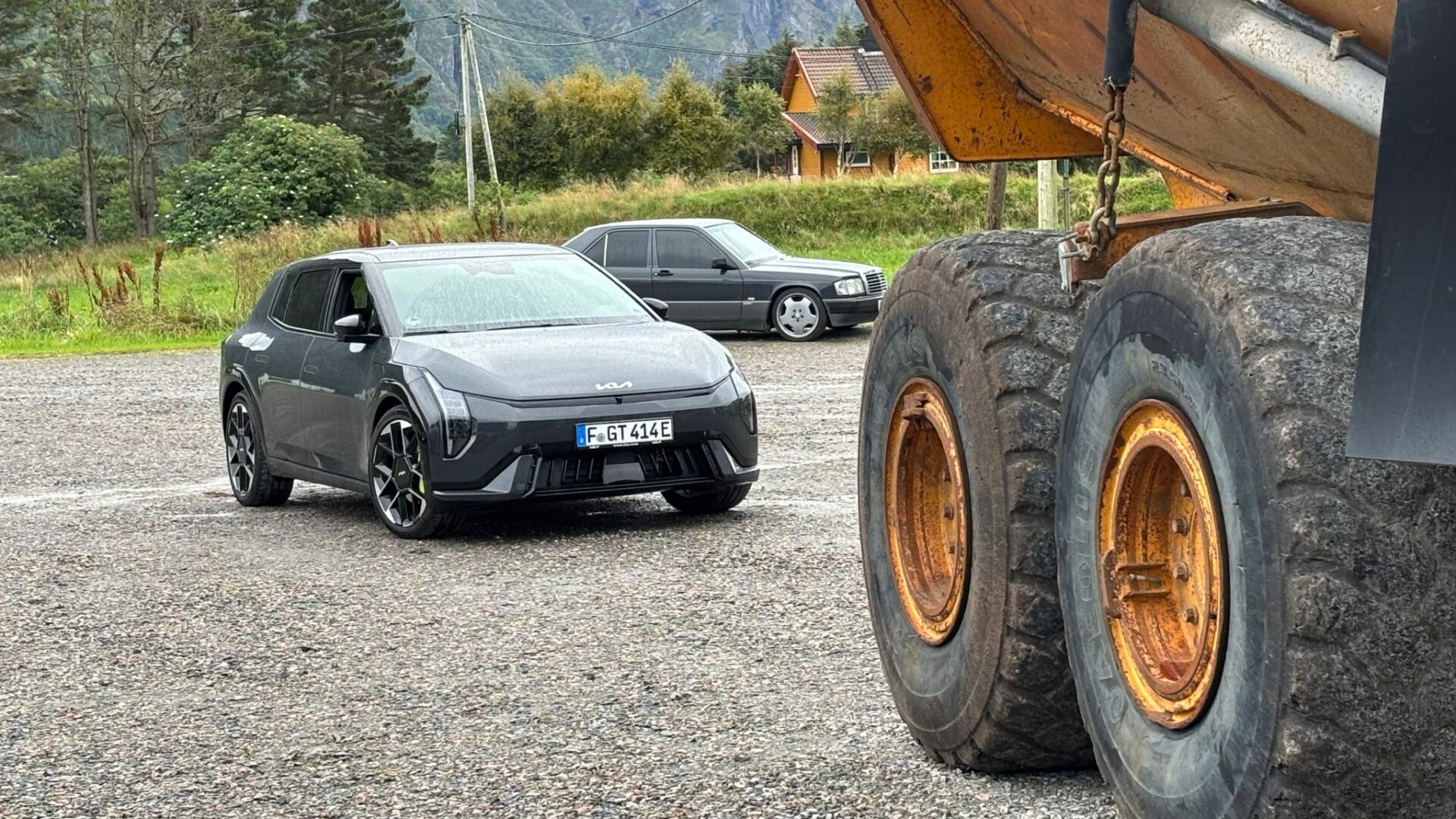 Vue de trois quarts avant de la nouvelle Kia EV4 GT grise stationnée sur un parking en gravier, avec les grosses roues d'un camion de chantier au premier plan à droite.