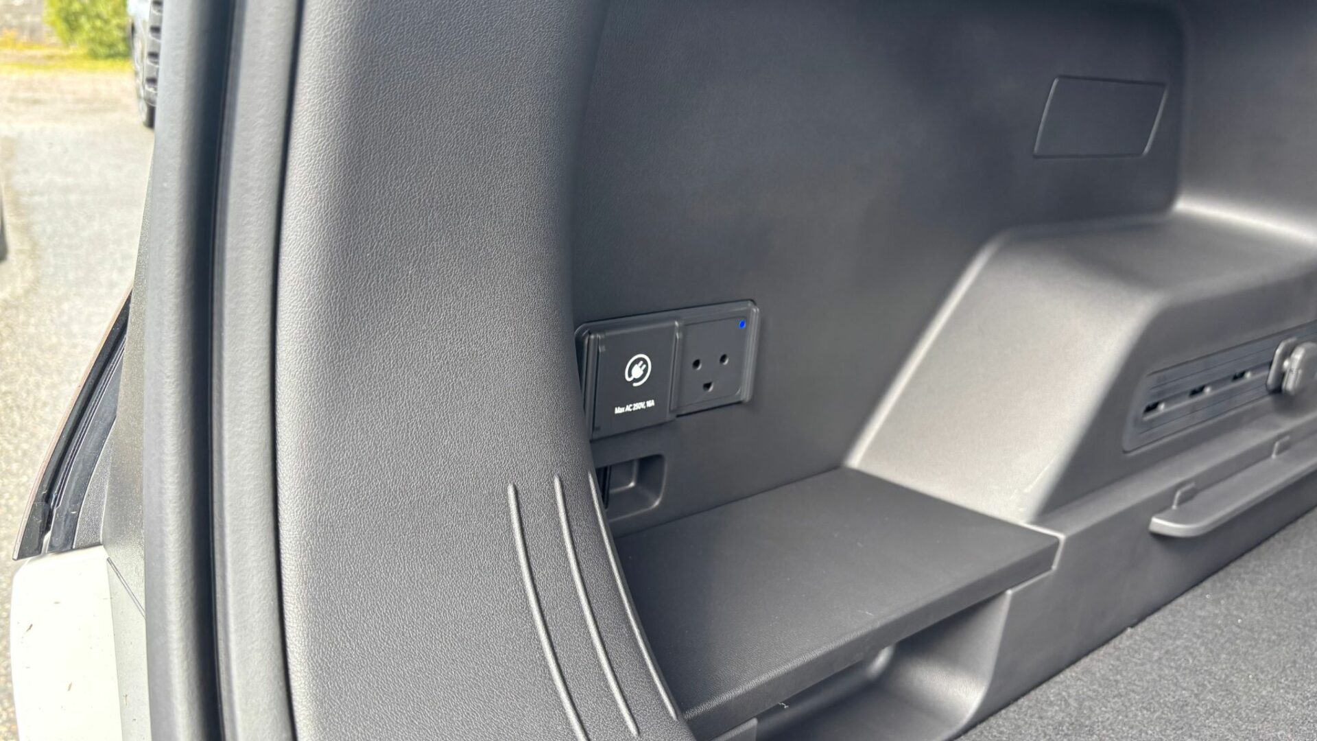 Gros plan sur la prise de courant domestique 230V intégrée dans l'habillage latéral du coffre de la Kia EV5 GT, illustrant la fonctionnalité Vehicle-to-Load (V2L).