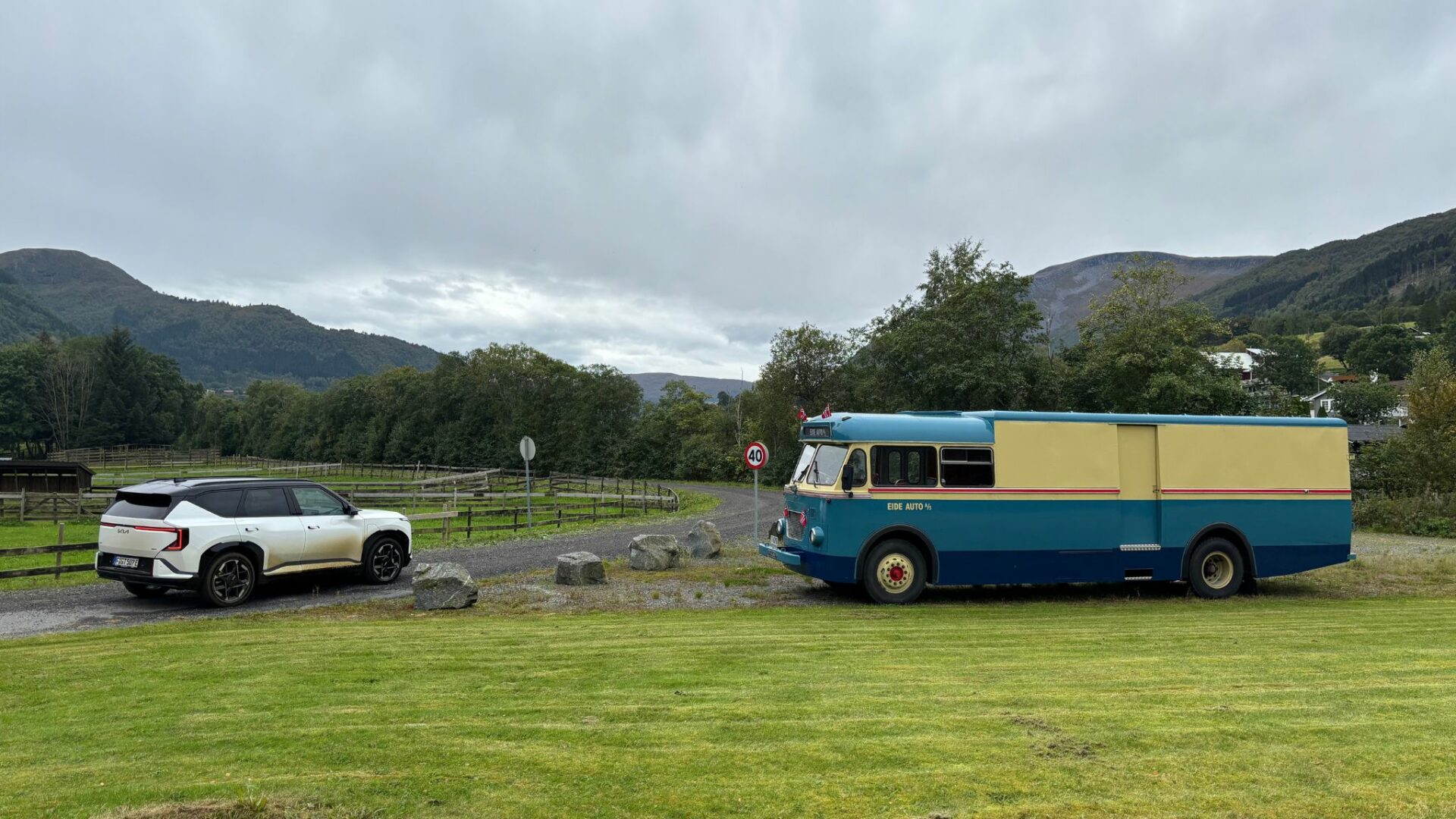 Vue d'ensemble de la nouvelle Kia EV5 GT blanche garée sur un chemin herbeux face à un ancien bus Volvo bleu et beige reconverti dans les paysages norvégiens.