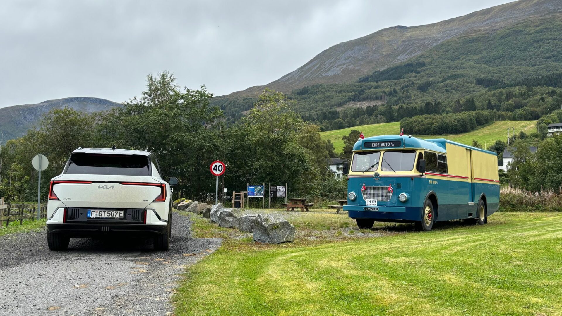 Vue de trois quarts arrière de la nouvelle Kia EV5 GT blanche garée sur un chemin de terre face à un bus Volvo vintage et aux montagnes en Norvège.