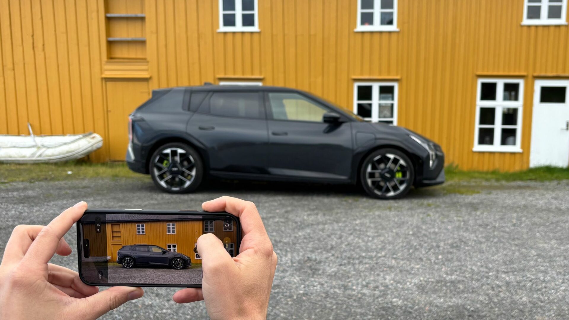 Un smartphone tenu par Mathias Dugenetay du blog-moteur.com capture en photo la nouvelle Kia EV4 GT grise stationnée de profil devant un bâtiment jaune en Norvège.
