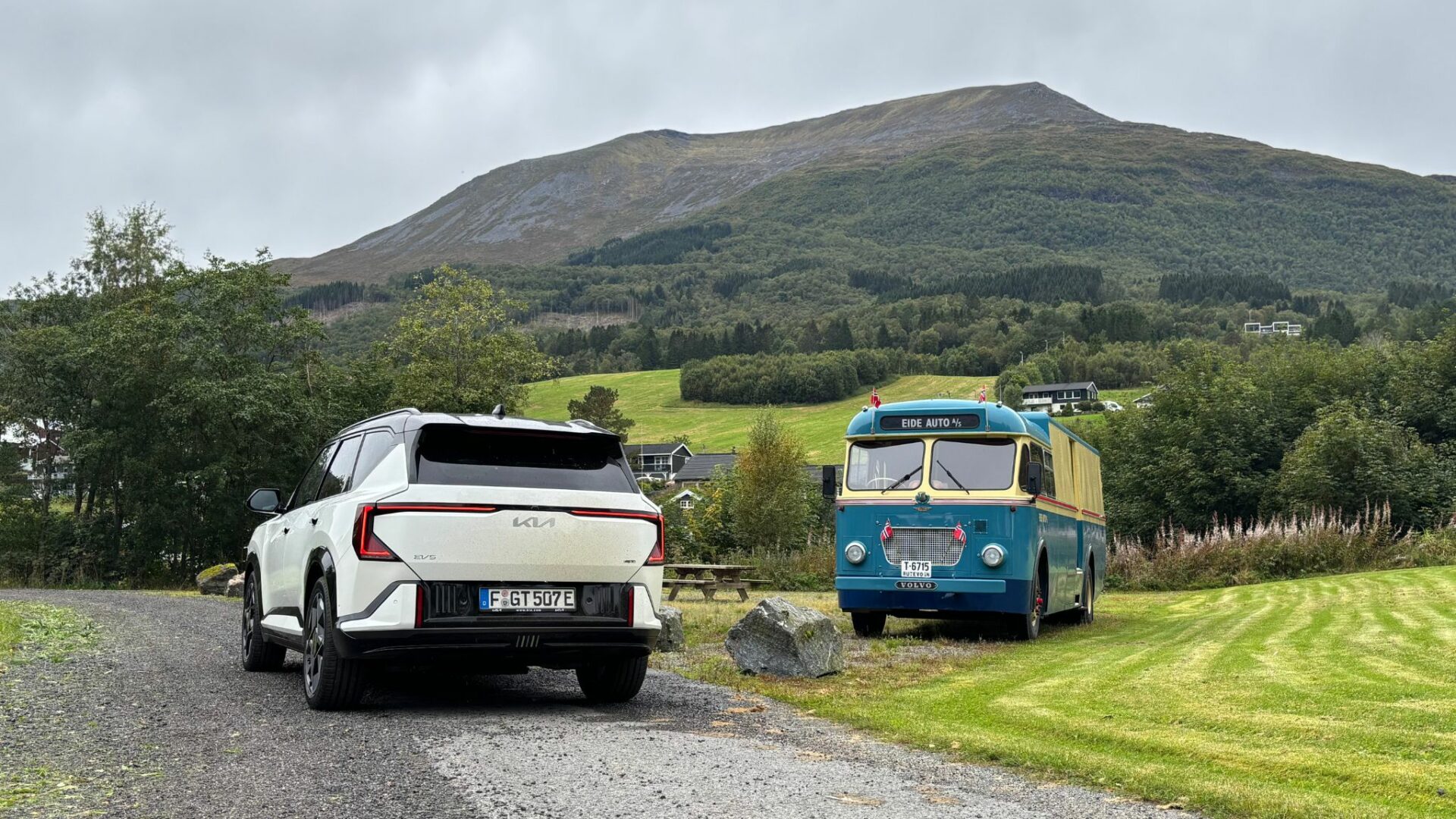 Vue de trois quarts arrière de la nouvelle Kia EV5 GT blanche garée sur un chemin de gravier, face à un ancien bus Volvo bleu et beige et à un grand sommet montagneux en Norvège.