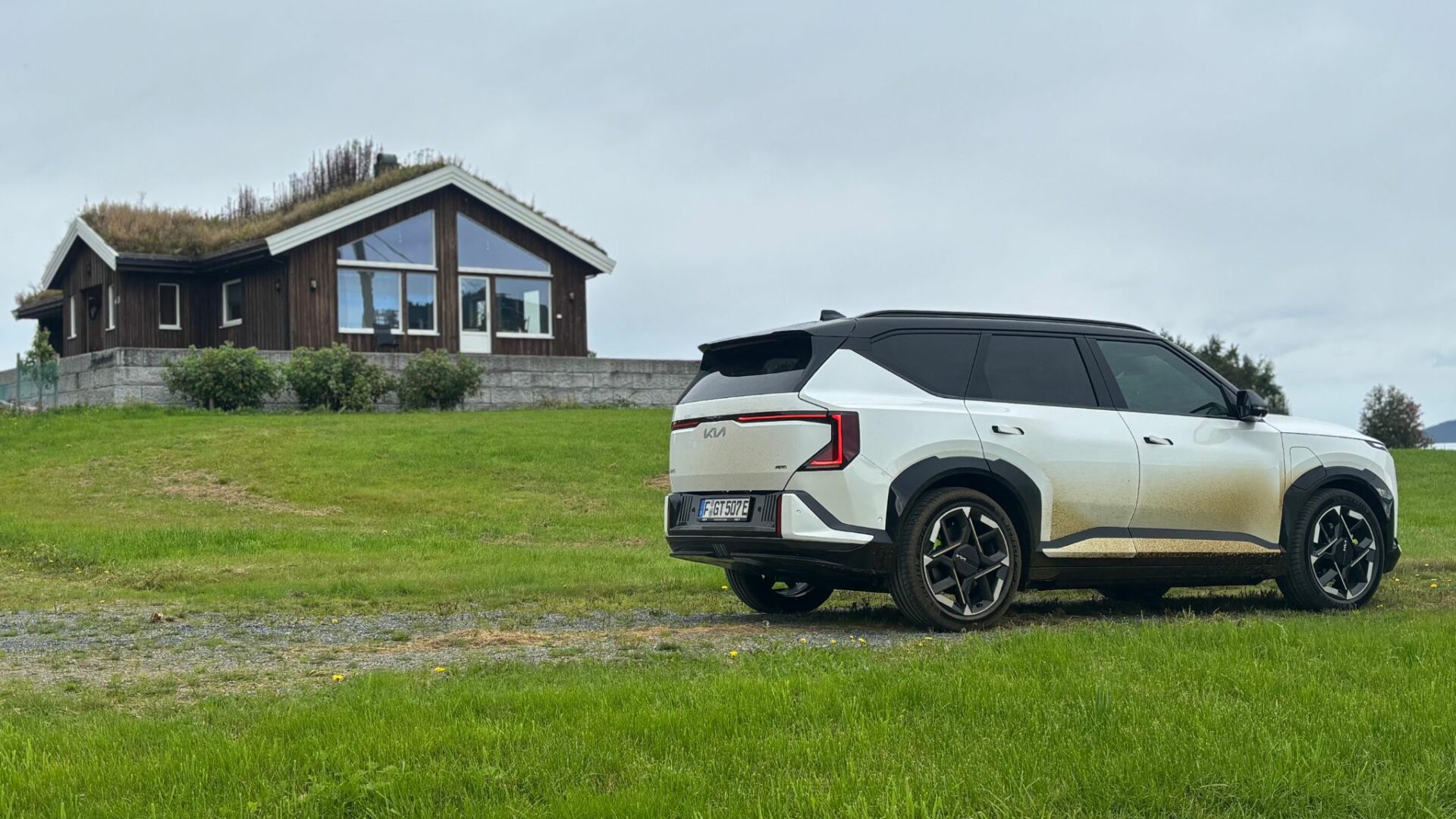 Vue de trois quarts arrière de la nouvelle Kia EV5 GT blanche, légèrement éclaboussée de boue, stationnée sur une prairie verdoyante devant une maison traditionnelle norvégienne au toit végétalisé.