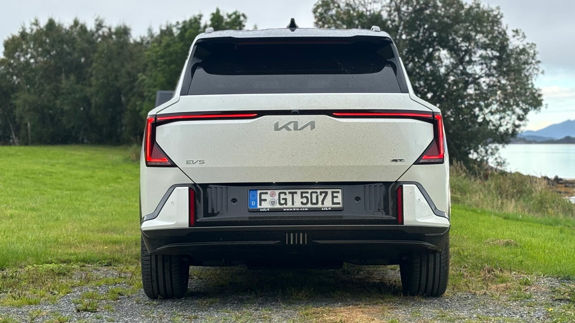 Vue arrière centrée de la nouvelle Kia EV5 GT blanche stationnée sur une piste en gravier bordant une pelouse verte et un plan d'eau en Norvège.