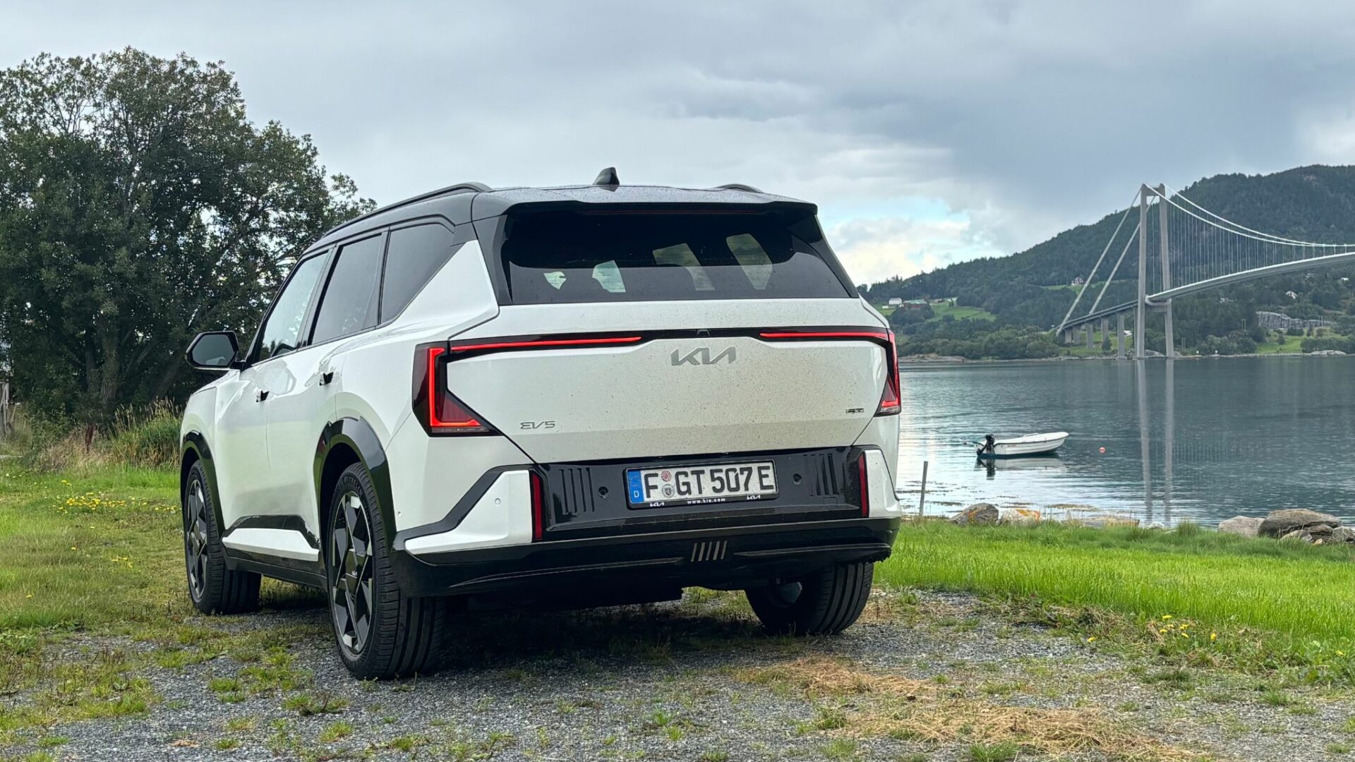 Vue de trois quarts arrière de la nouvelle Kia EV5 GT blanche stationnée sur un chemin côtier avec un fjord, un bateau et un pont suspendu en arrière-plan en Norvège.