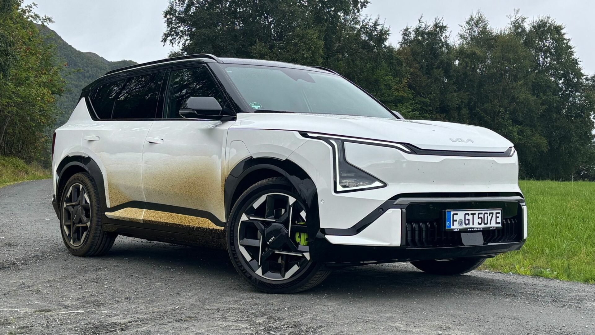 Vue de trois quarts avant de la nouvelle Kia EV5 GT blanche, légèrement éclaboussée de boue sur les flancs, stationnée sur un chemin de terre bordant une prairie et des arbres en Norvège.