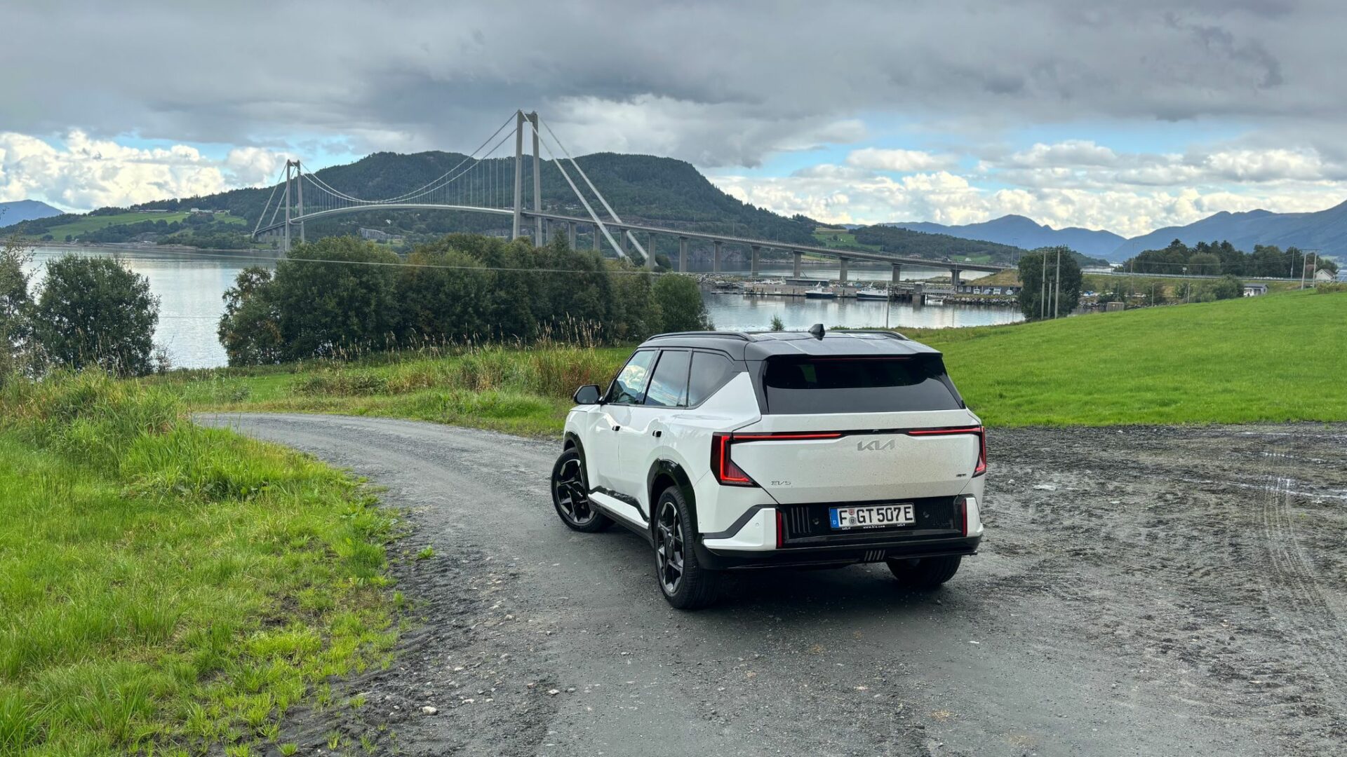 Vue large de trois quarts arrière de la nouvelle Kia EV5 GT blanche arrêtée sur un sentier en gravier dominant un fjord, avec un grand pont suspendu et des montagnes en arrière-plan en Norvège.