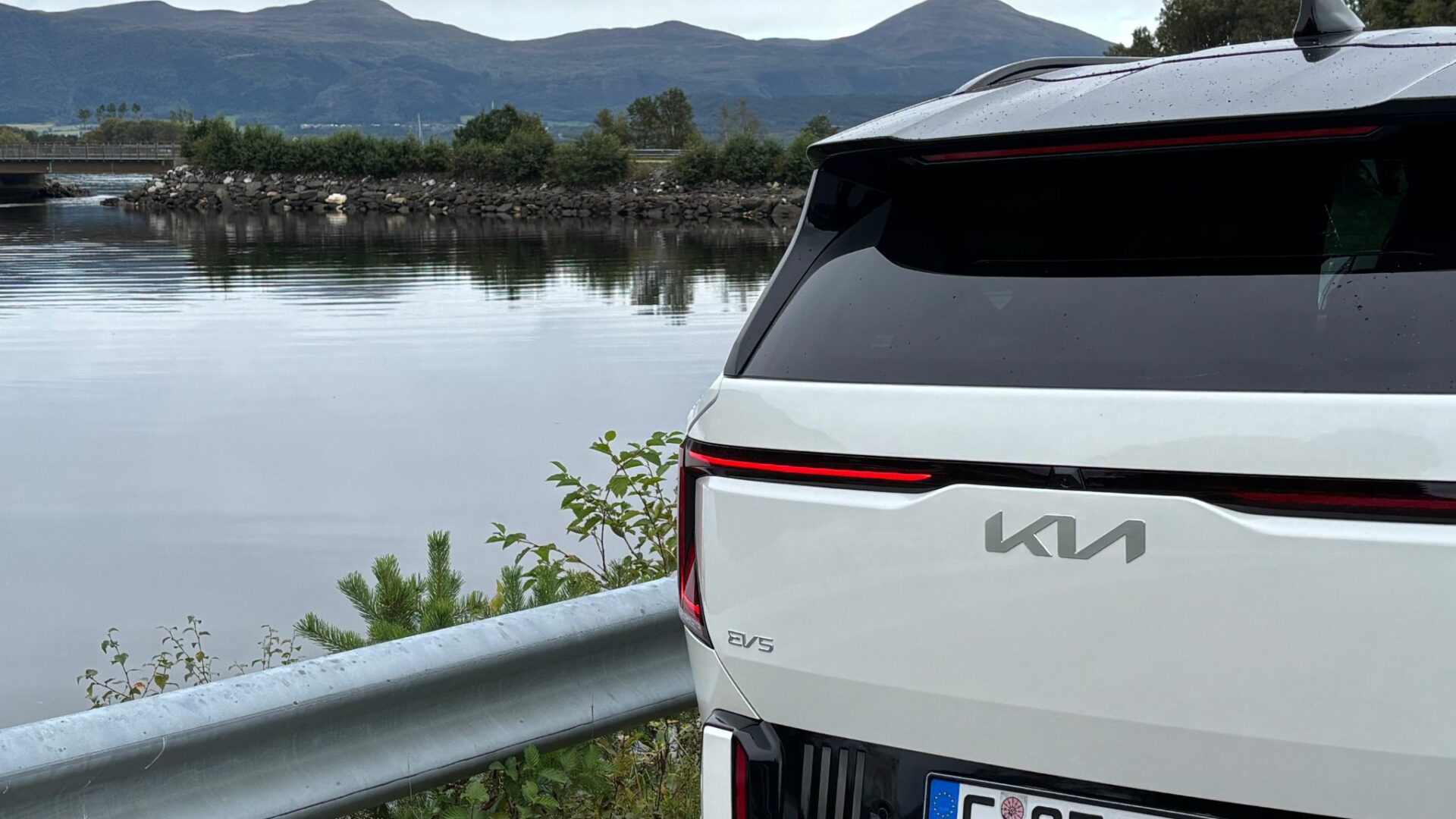 Gros plan sur la partie arrière de la nouvelle Kia EV5 GT blanche, mettant en valeur son bandeau lumineux et ses monogrammes, garée au bord d'un plan d'eau et de montagnes en Norvège.