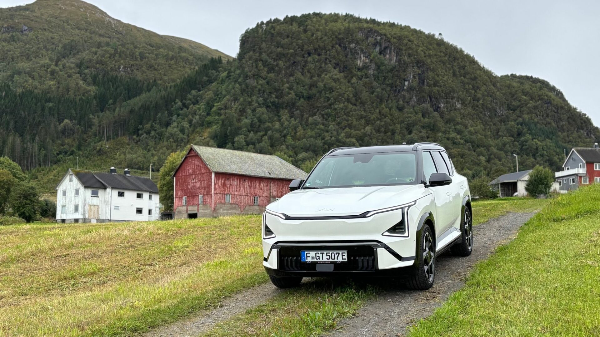 Vue de face de la nouvelle Kia EV5 GT blanche stationnée sur un chemin de terre sinuant à travers des prairies, avec des granges traditionnelles rouges et blanches et des montagnes verdoyantes en Norvège.