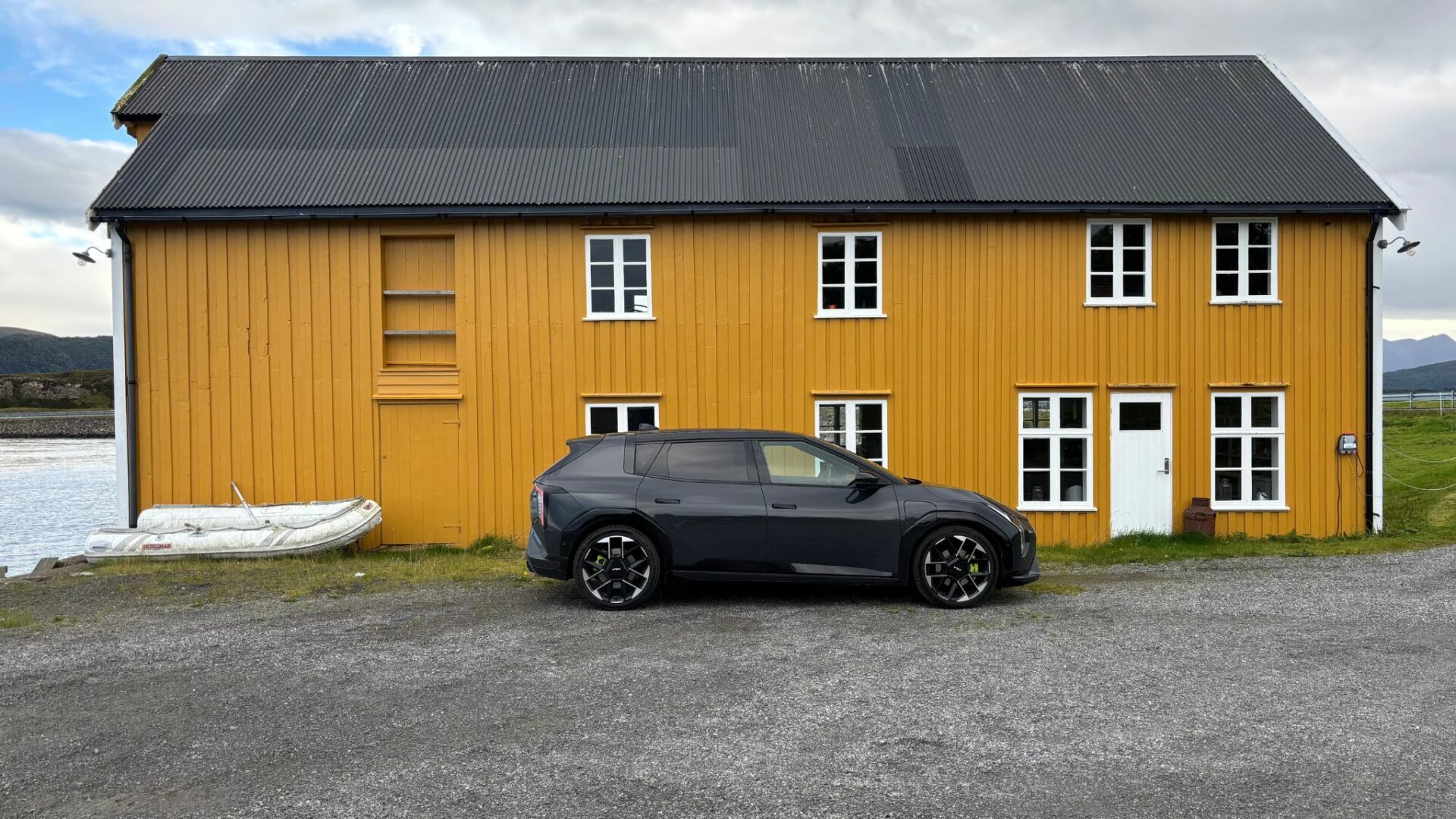 Vue de profil de la nouvelle Kia EV4 GT grise stationnée sur un chemin de gravier devant un grand bâtiment en bois jaune typique de Norvège.