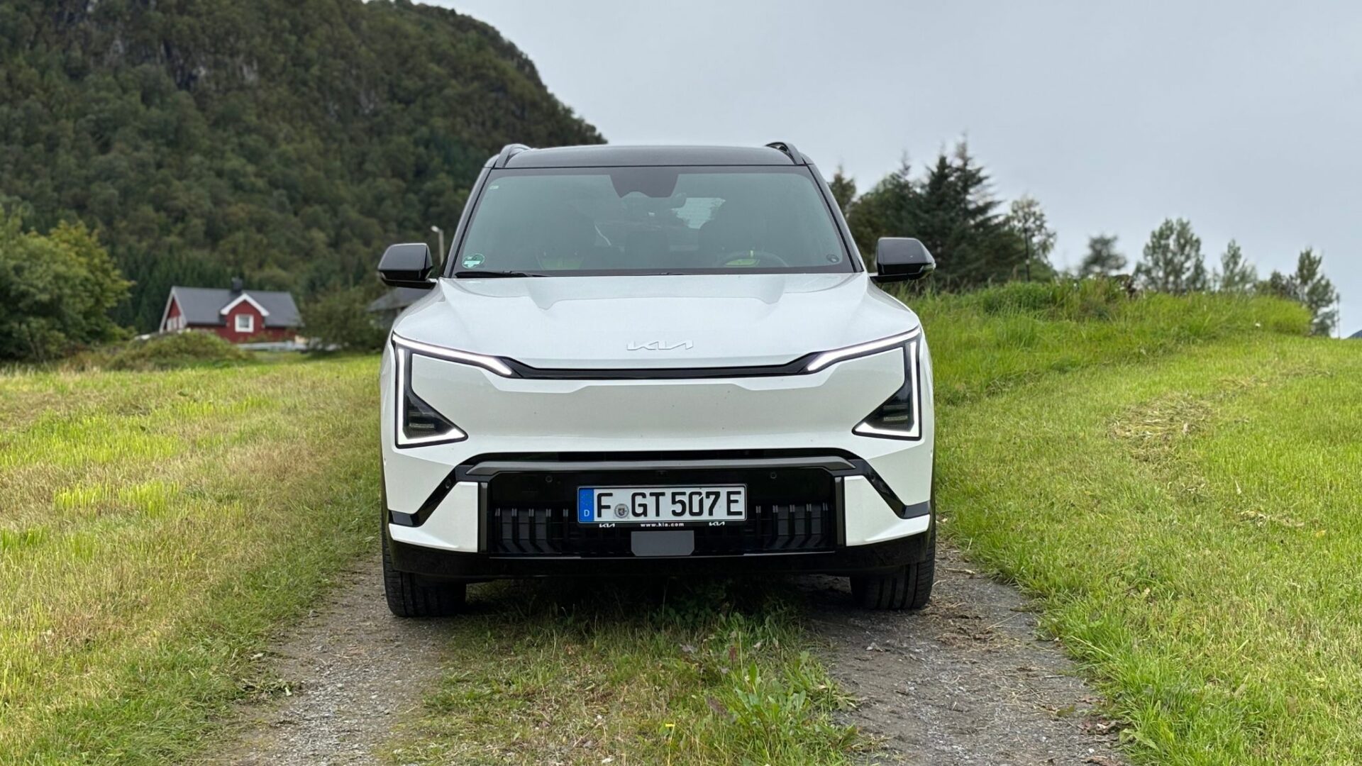Vue frontale centrée de la nouvelle Kia EV5 GT blanche stationnée sur un chemin de terre bordé de prairies verdoyantes et d'une maison traditionnelle en Norvège.