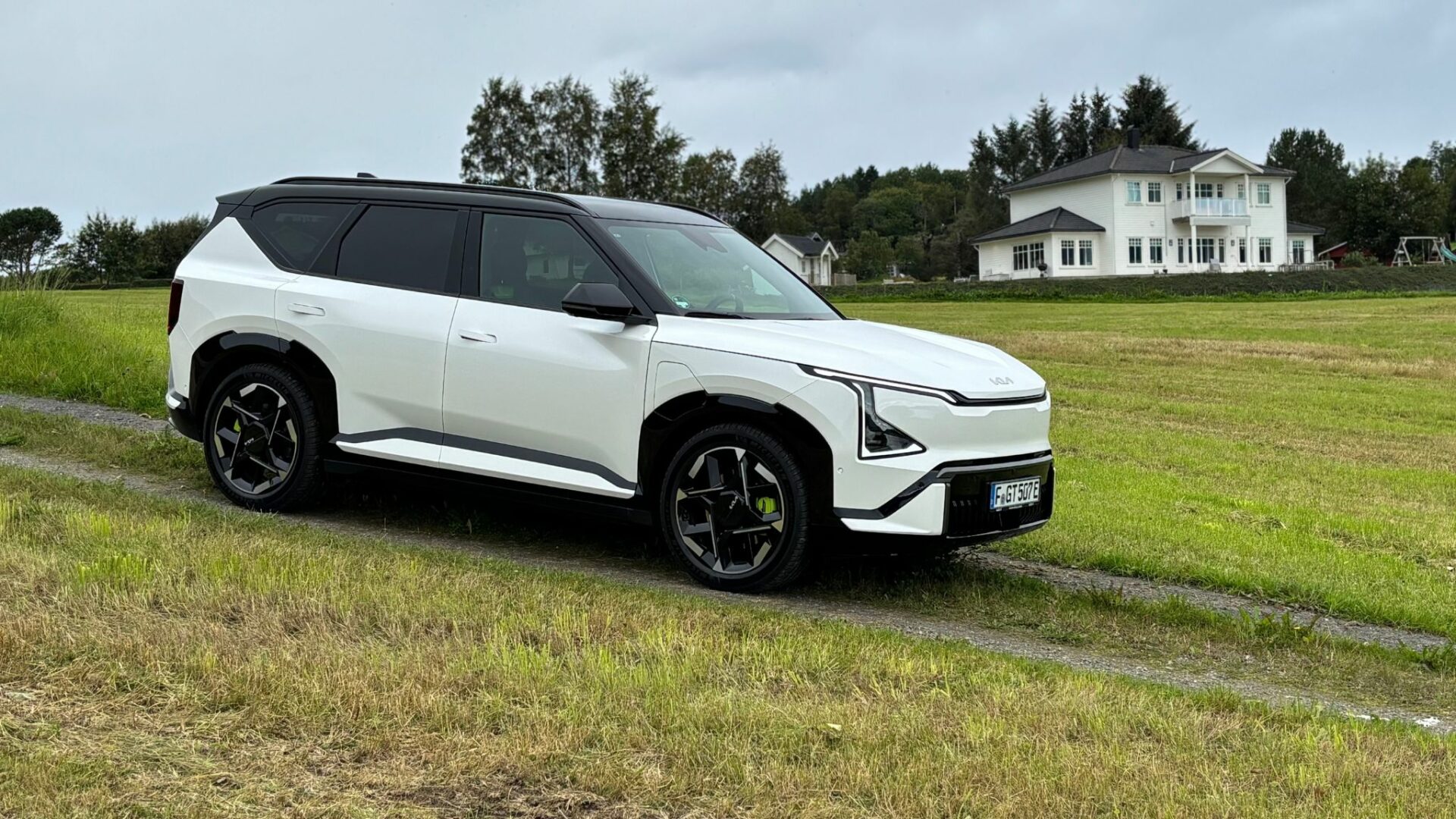 Vue de trois quarts avant de la nouvelle Kia EV5 GT blanche stationnée sur un chemin herbeux avec une grande maison blanche en arrière-plan en Norvège.