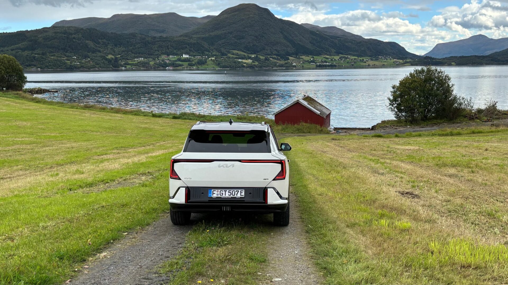 Vue arrière centrée de la nouvelle Kia EV5 GT blanche garée sur un chemin de terre descendant vers un fjord, avec une cabane rouge et des montagnes en arrière-plan en Norvège.