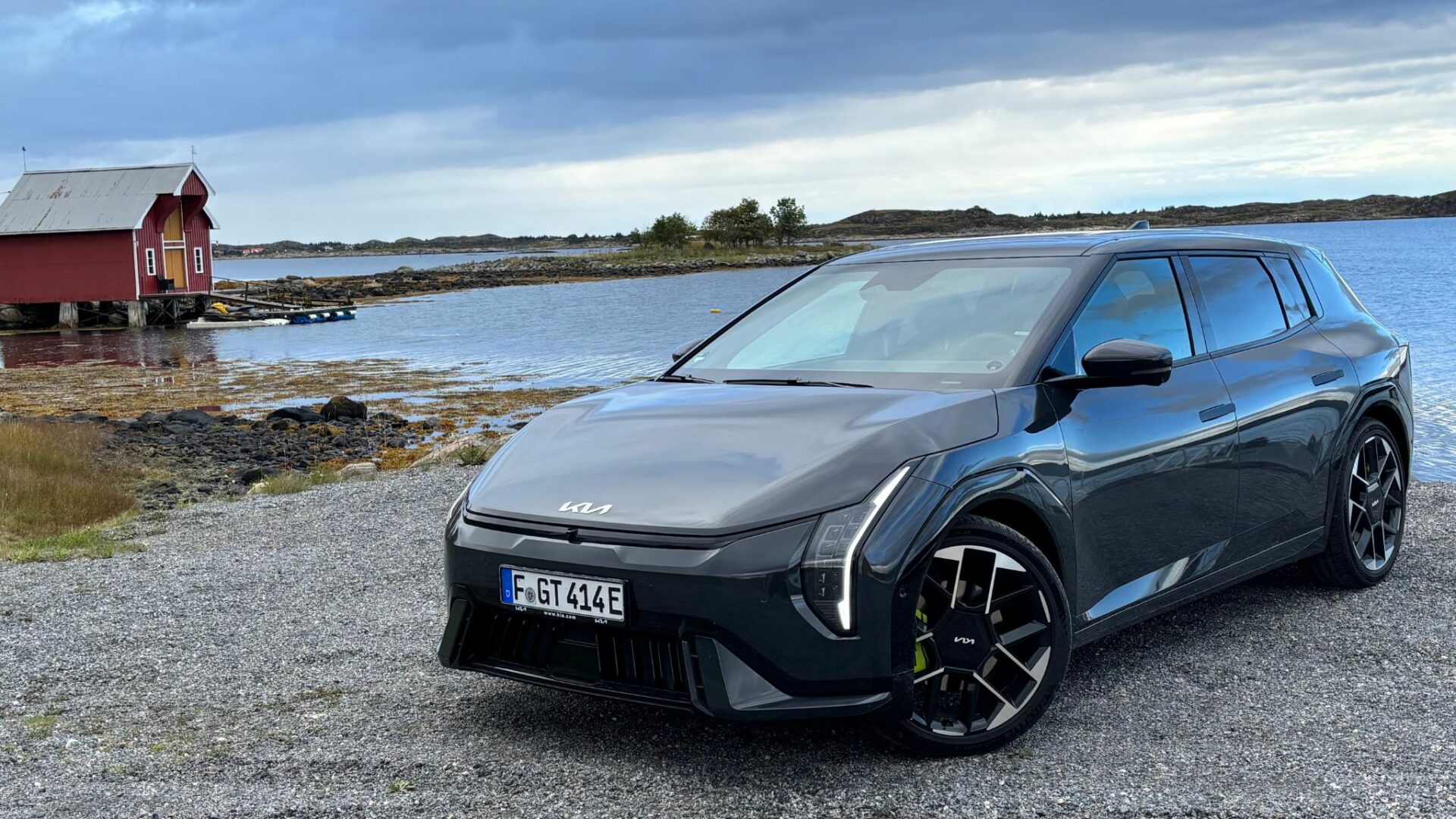 Vue de trois quarts avant de la nouvelle Kia EV4 GT grise stationnée au bord de l'eau devant une cabane traditionnelle rouge en Norvège.