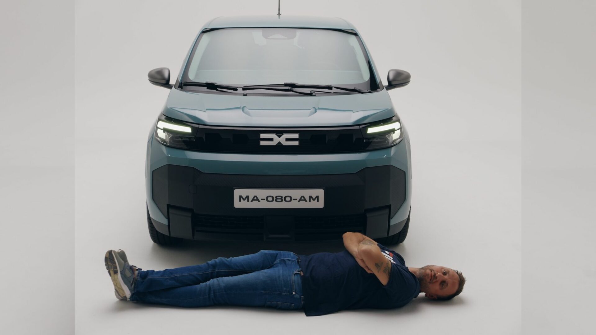 Le célèbre youtubeur automobile Victor Diakov allongé en travers devant la face avant de la Nouvelle Dacia Spring pour illustrer ses 18 cm de largeur gagnés.