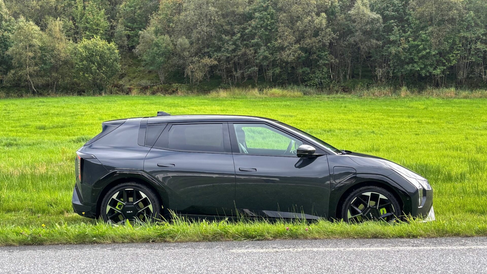 Vue de profil de la nouvelle Kia EV4 GT grise garée le long d'une route devant un vaste champ verdoyant et une lisière de forêt en Norvège.